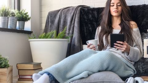 Wellness and Reading Woman with ebook and cup of tea on couch in blanket self care
